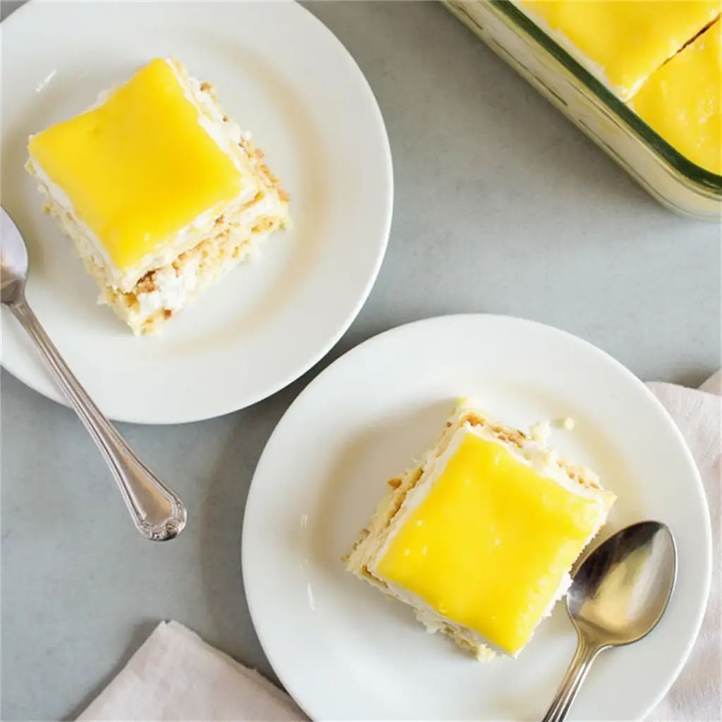 Two slices of limoncello tiramisu served on white plates, showcasing layers of mascarpone, biscuit, and lemon curd, with a baking dish in the background.