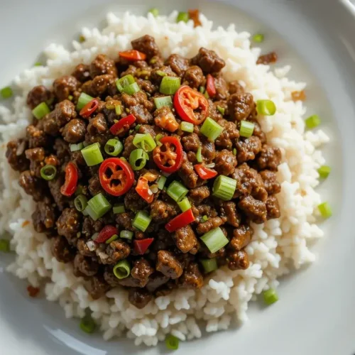 A plate of Mongolian ground beef recipe served over white rice, topped with fresh green onions and red chili peppers, showcasing a flavorful and comforting dish.